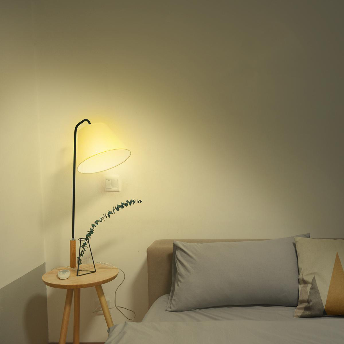 Eine Lampe mit beigem Schirm steht auf einem kleinen Tisch mit einer Vase und einer Pflanze. Im Hintergrund befindet sich ein Bett.