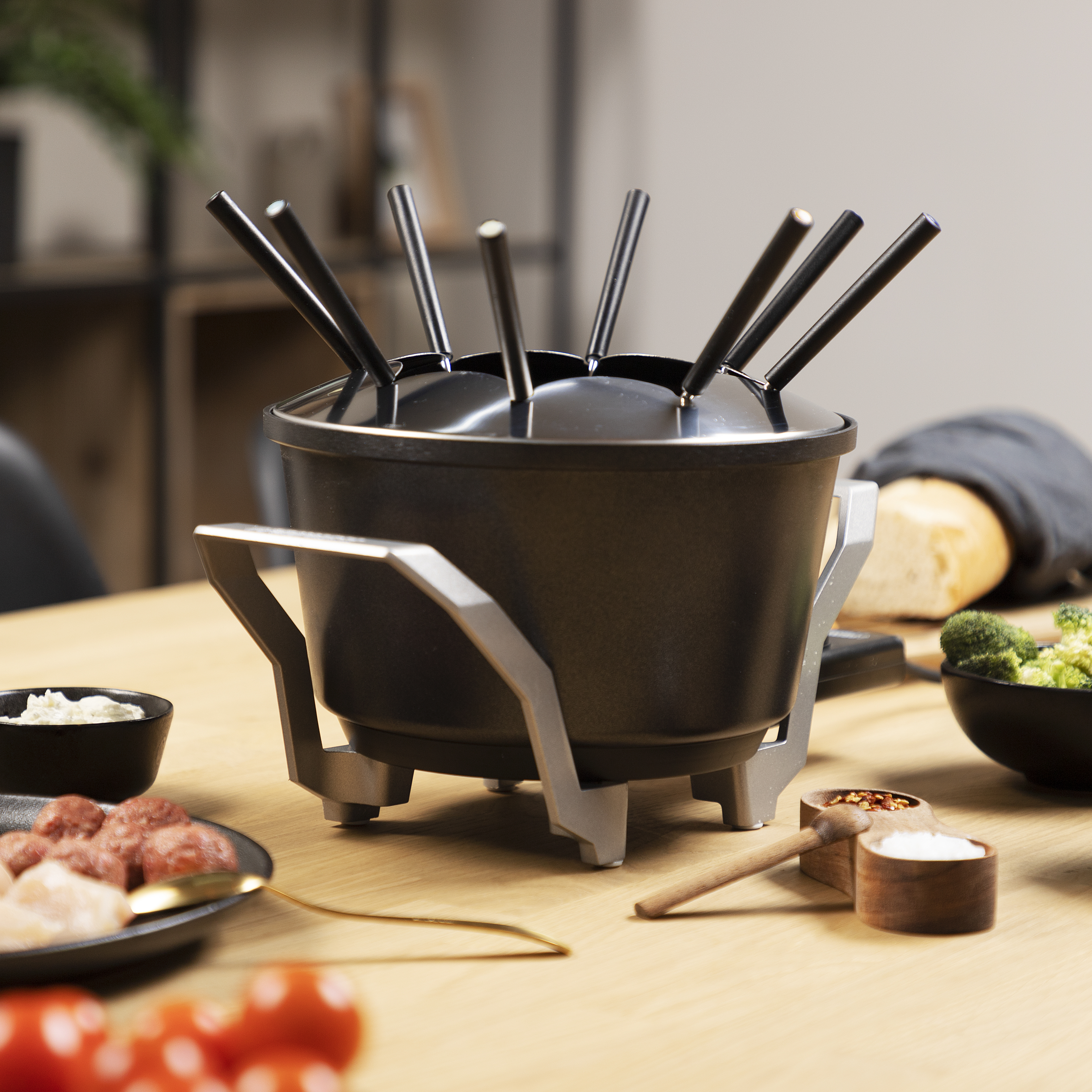 A black fondue pot with forks, food, and seasonings on a wooden table. A white bowl of sauce is present.