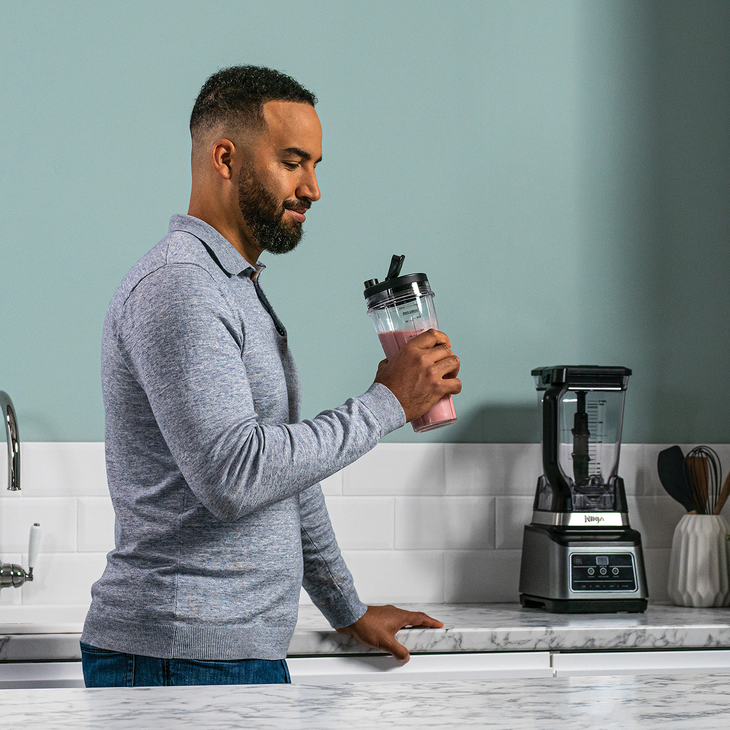 Man houdt een smoothiebeker vast naast een blender. De achtergrond is een lichtblauwe muur.