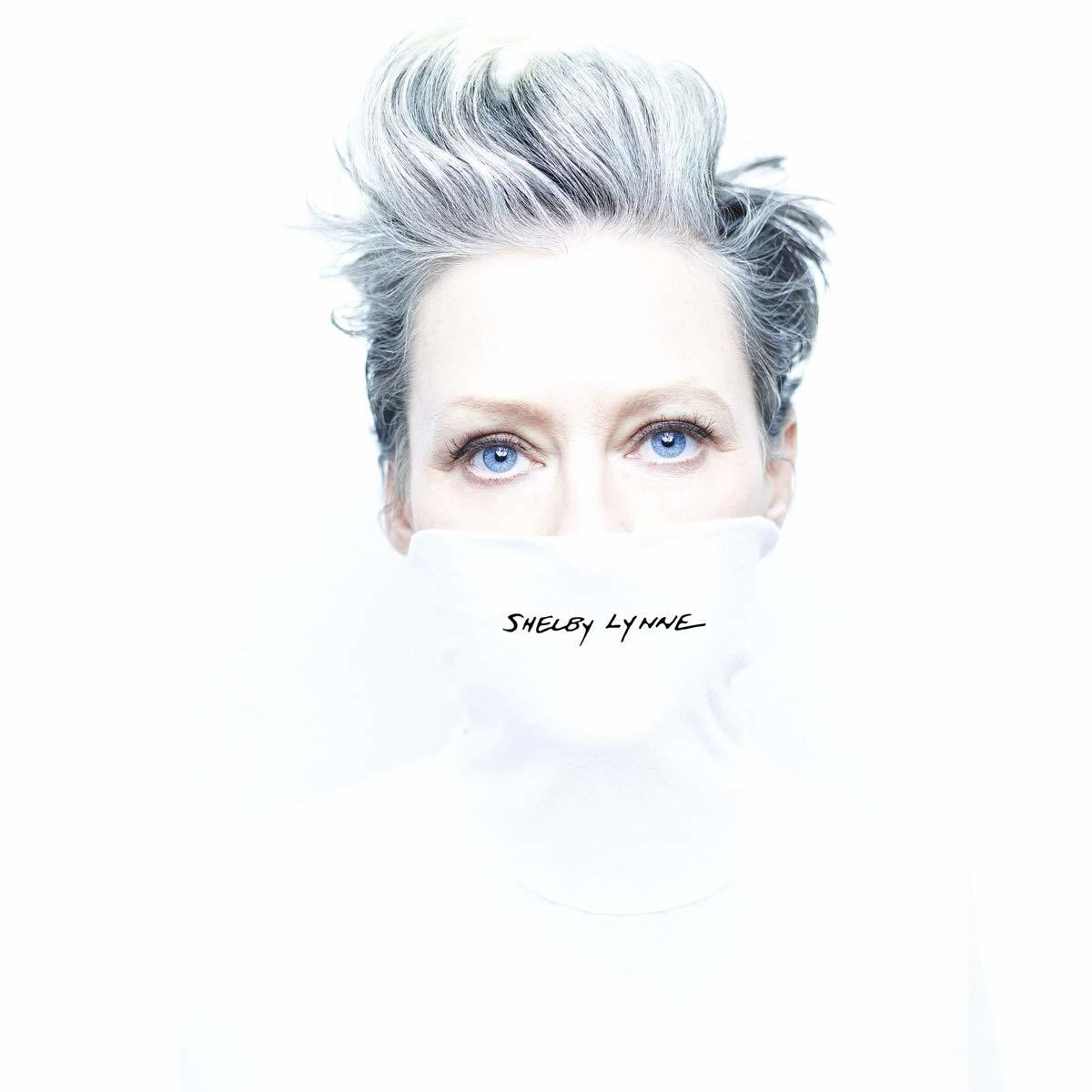 Egy kék szemű, fehér hajú nő fehér kendőt visel a szája előtt. A "Shelby Lynne" név van ráírva.