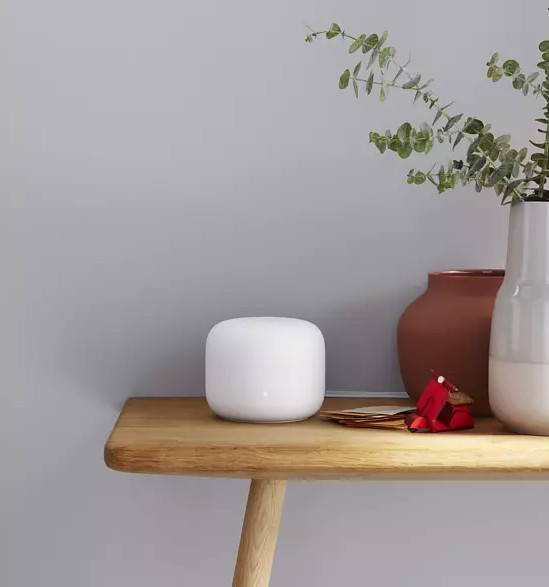 Google Nest Wifi blanc sur une table en bois. Vase, plante et modèle de scooter rouge.