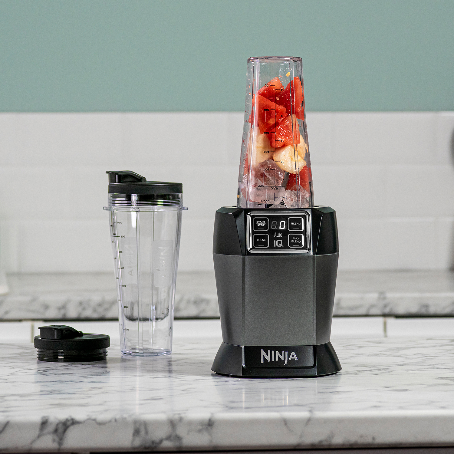A blender with strawberries and fruit, a clear cup, and a black lid sits on marble.