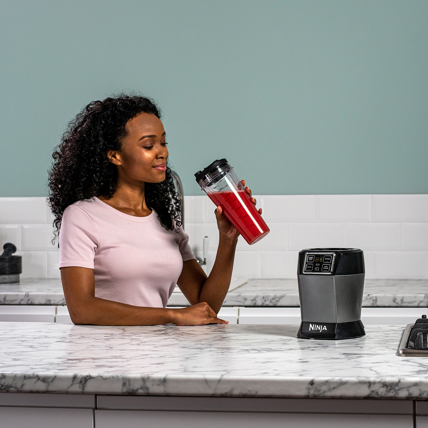 Woman with a pink shirt holding a red blender cup, other items are on the table.