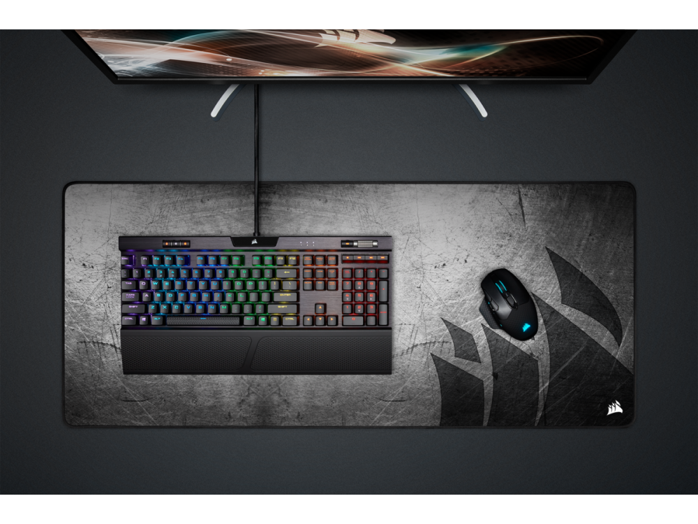 Teclado, ratón y monitor en una alfombrilla gris, logo Corsair visible.