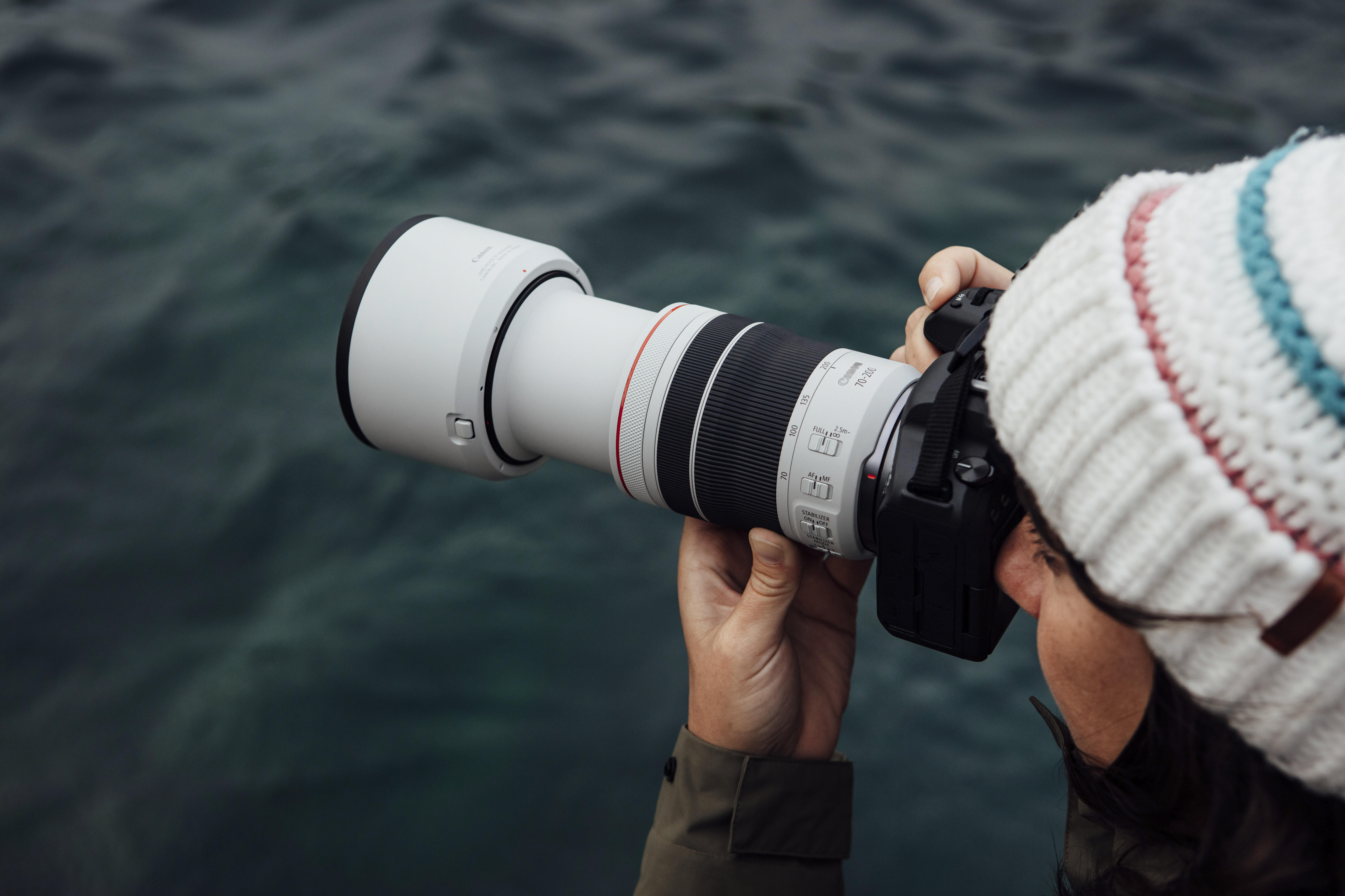 Eine Person hält eine Kamera mit einem langen weißen Objektiv und fotografiert Wasser. Sie trägt eine Strickmütze.