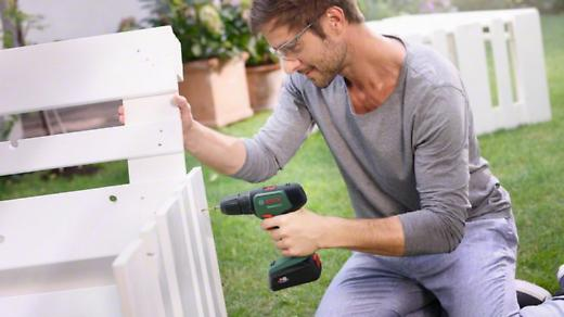 Férfi zöld-fekete fúróval fehér padot szerel össze.