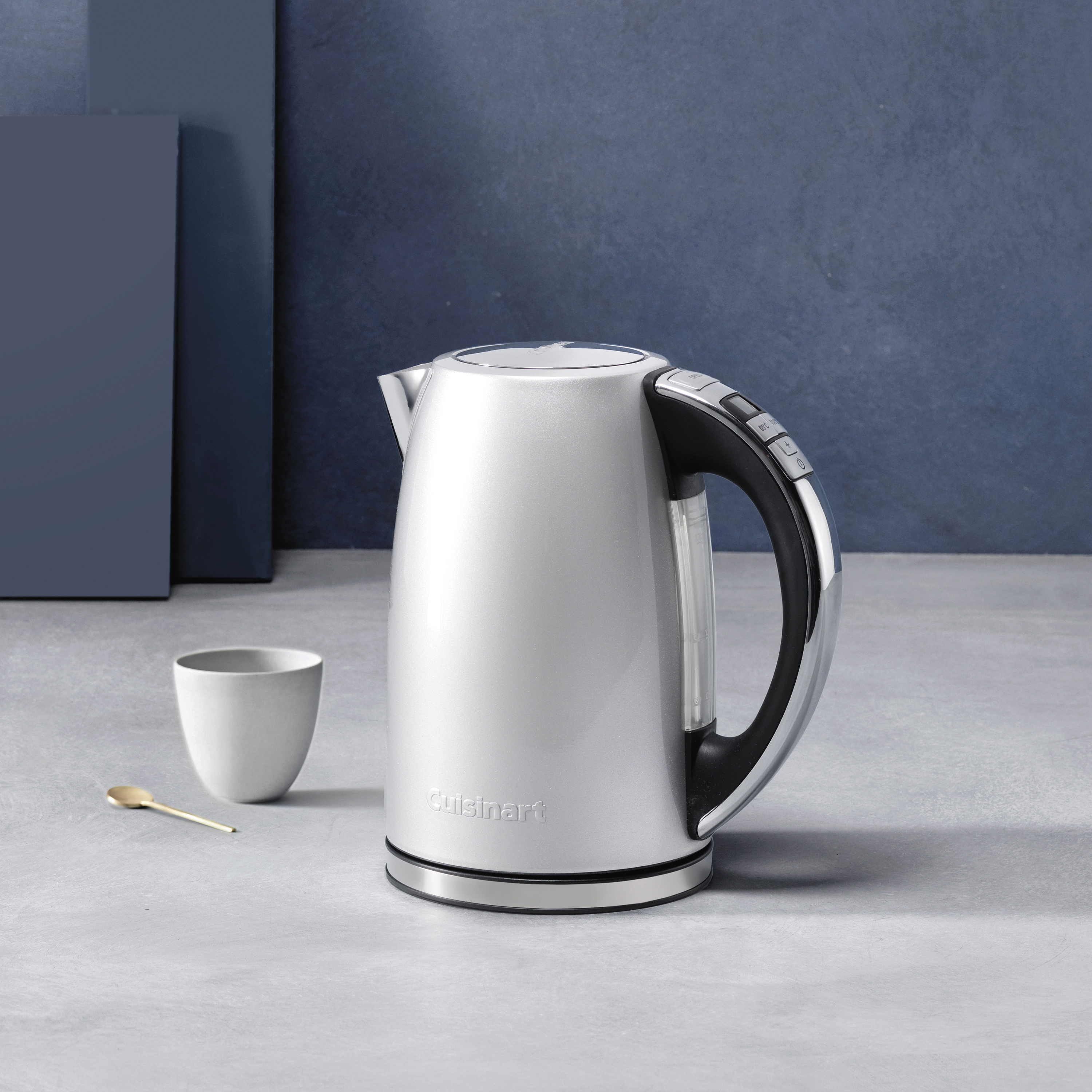 Bouilloire électrique argentée avec poignée noire et tasse blanche à côté. Comptoir gris clair, fond bleu et gris.