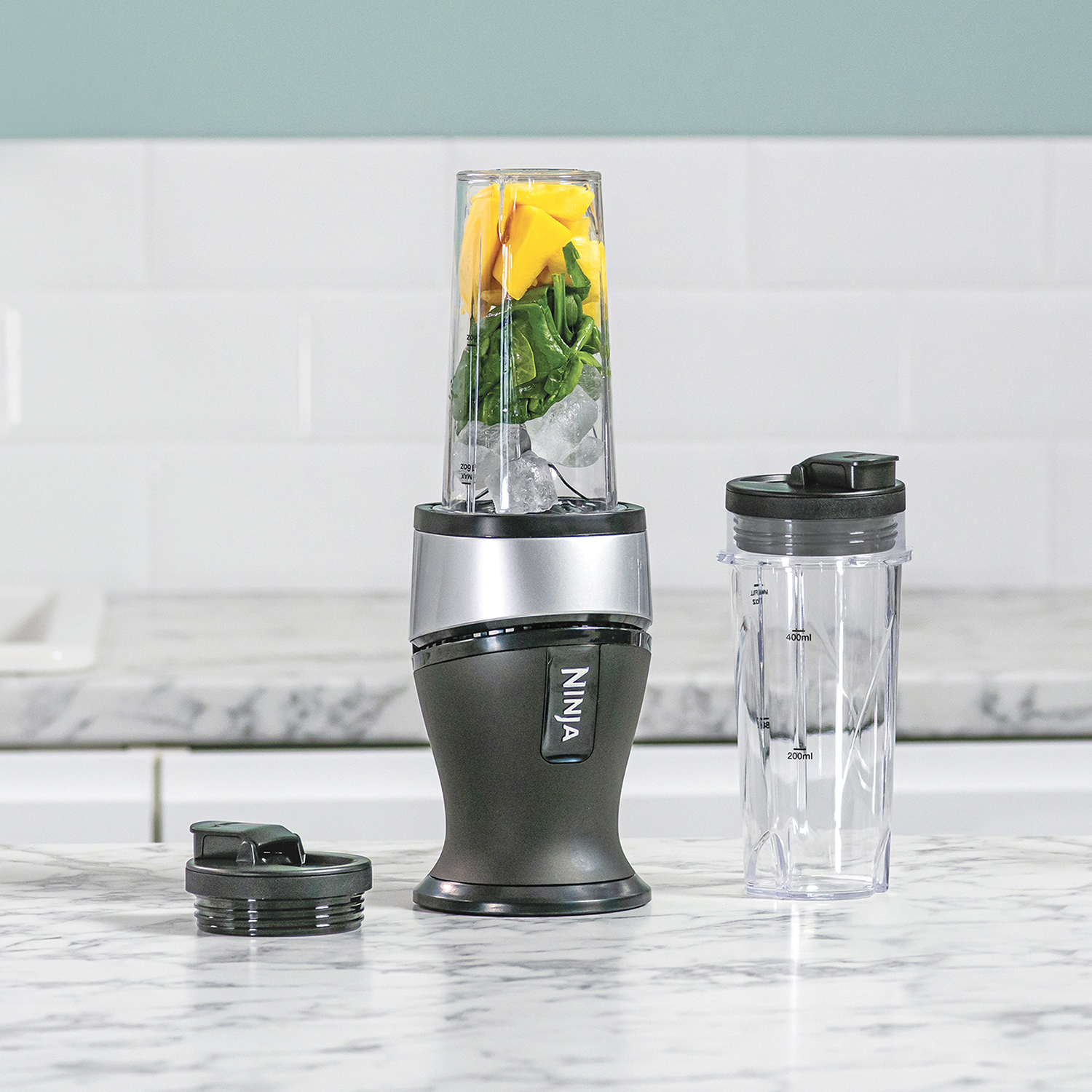A blender with fruits and ice. Another container and lid are next to it. White background.