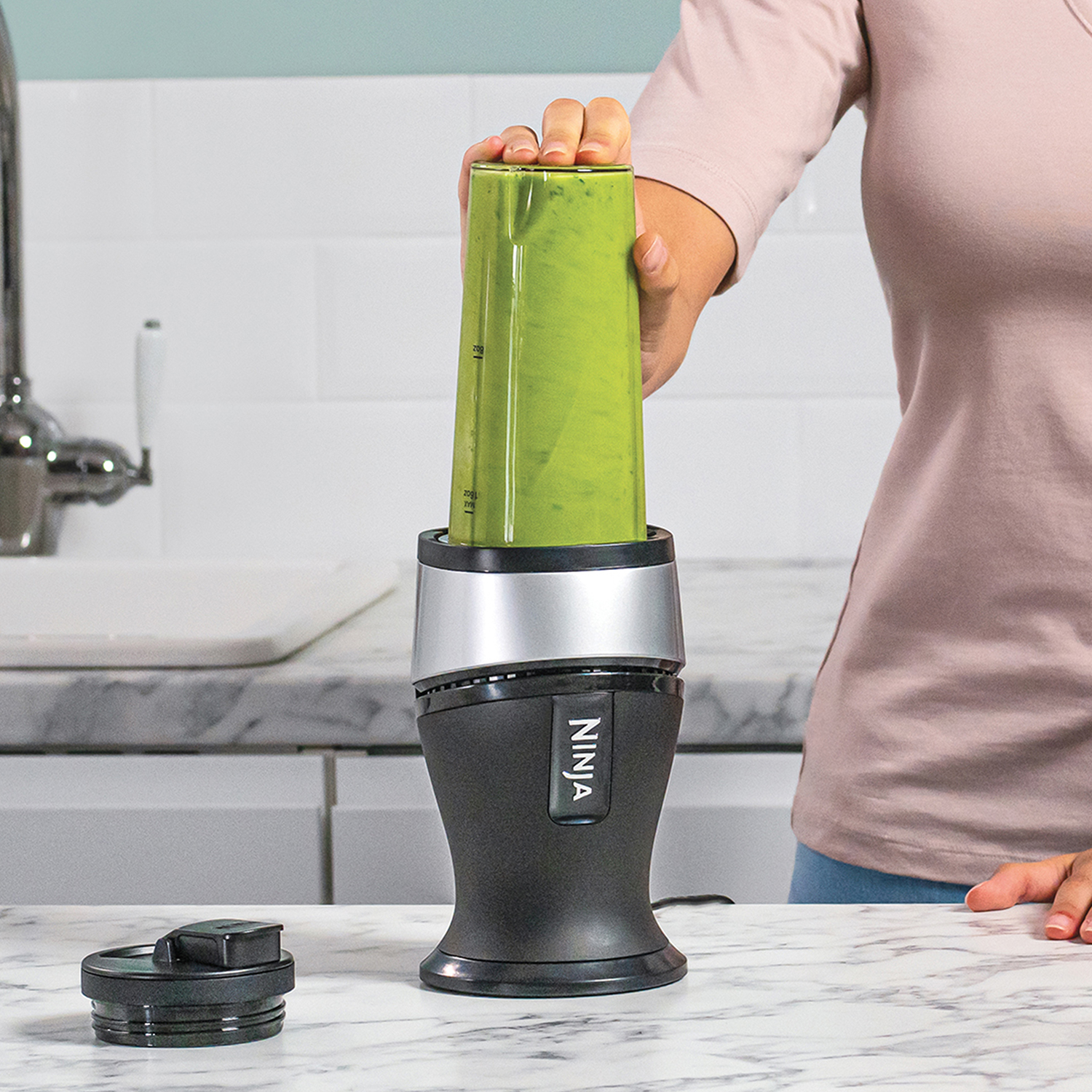 A woman blending a green smoothie in a black and silver Ninja blender. Marble countertop.