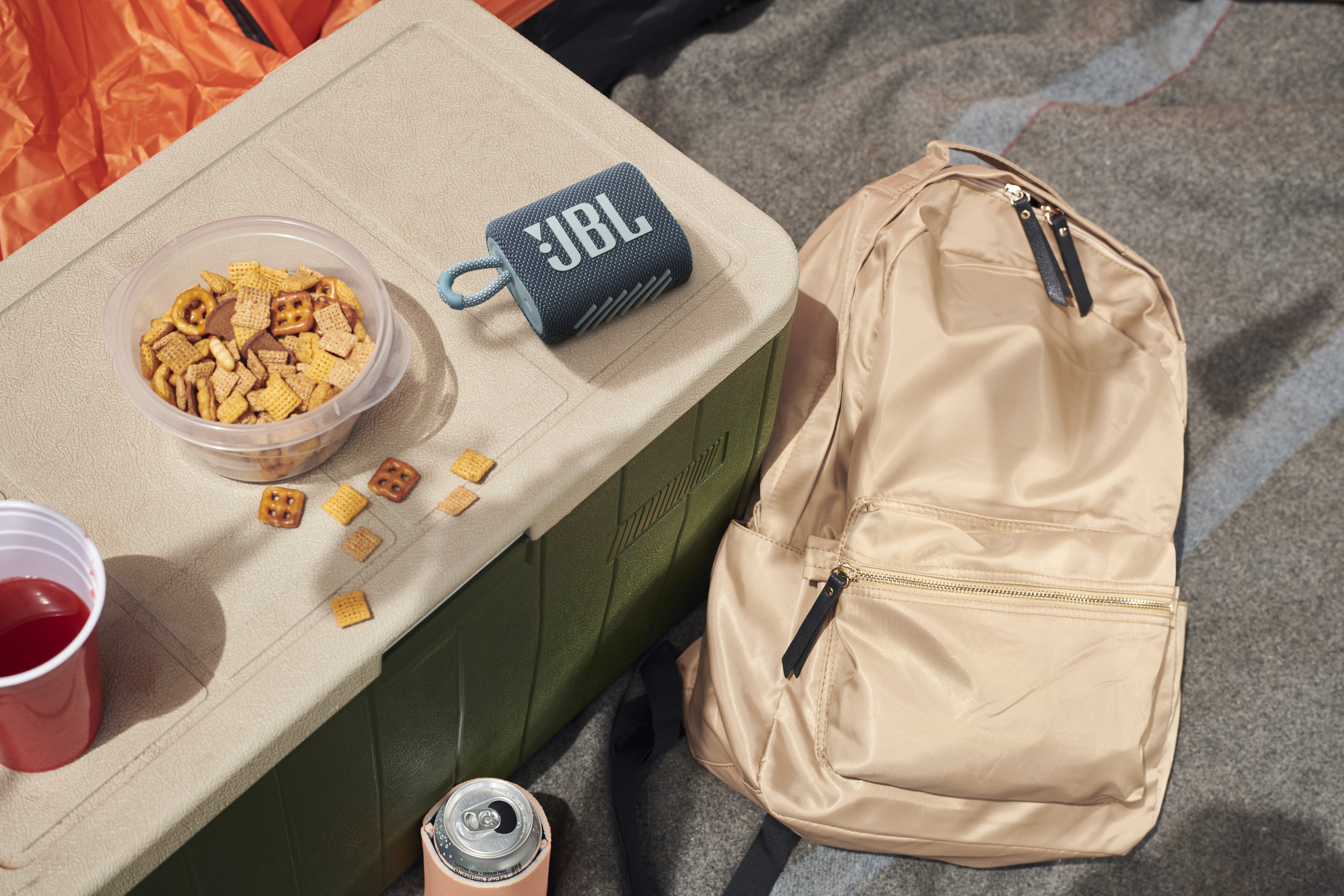 Szene im Freien mit einem JBL-Lautsprecher, Snacks und einem Rucksack auf einer Decke, neben einer Kühlbox.