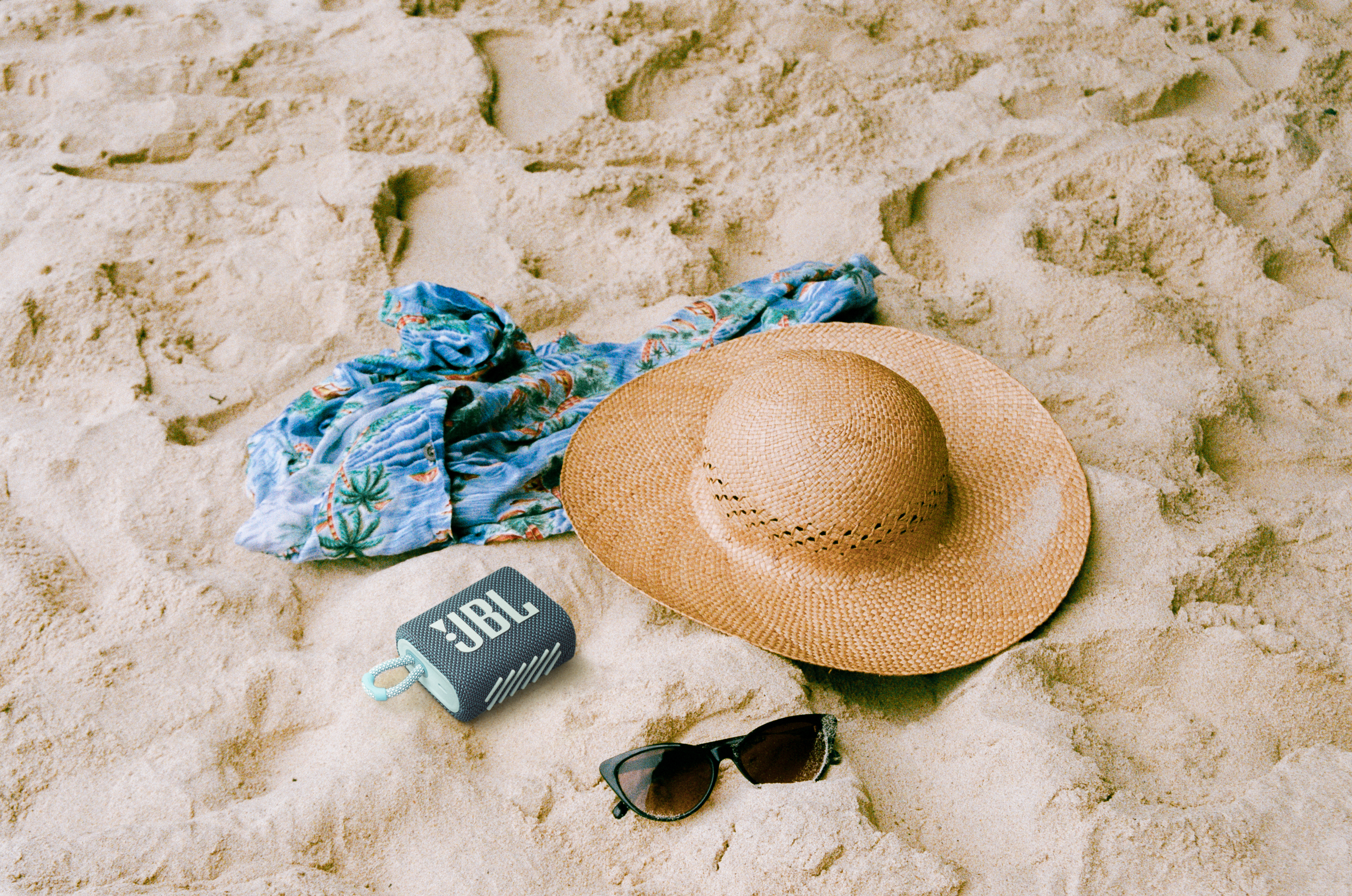 Strandszene mit Hut, Sonnenbrille, Lautsprecher und gemustertem Hemd auf Sand.