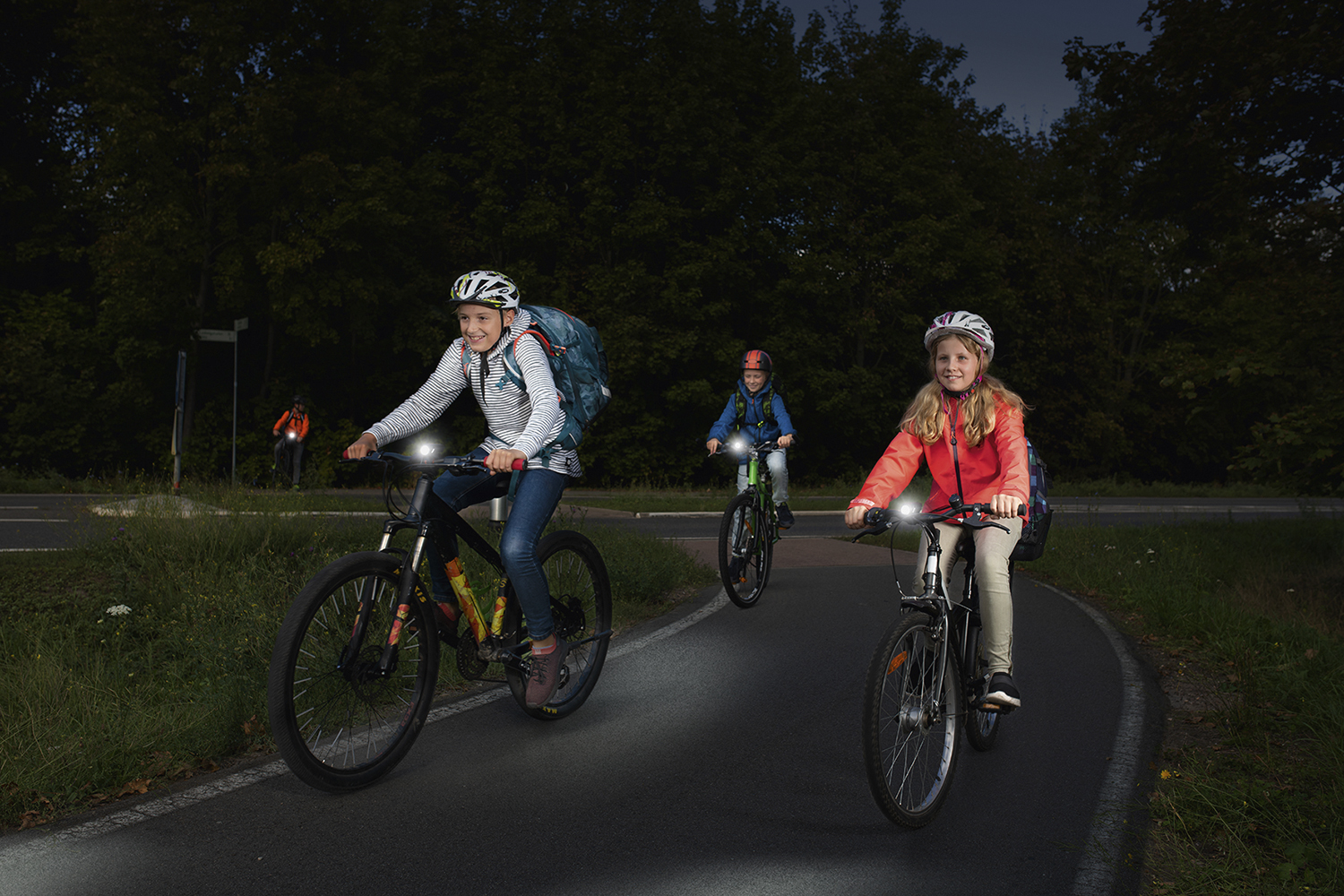 Kinder fahren nachts Rad. Fahrräder haben Lichter. Bäume und eine Straße im Hintergrund.