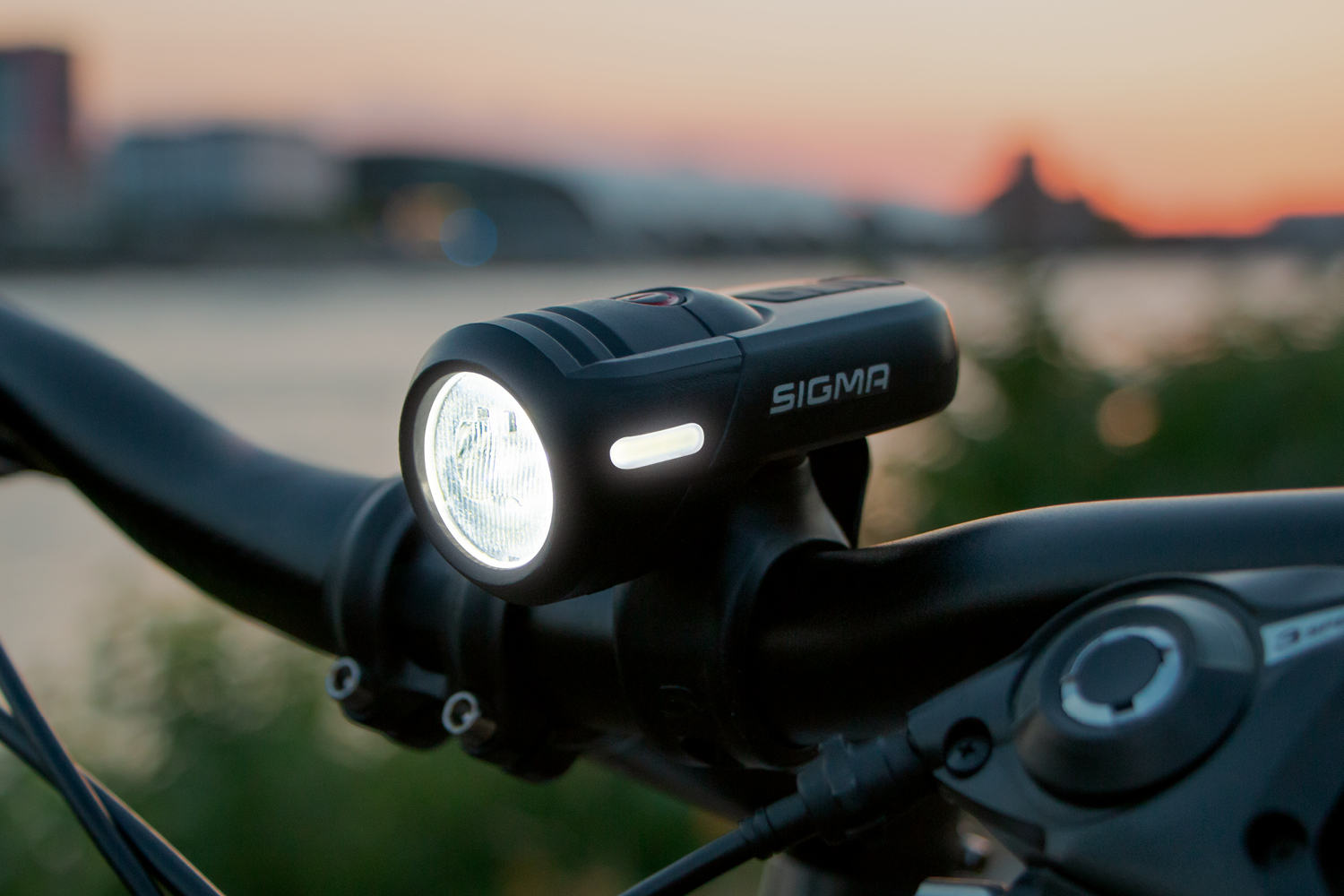 Fahrradscheinwerfer, schwarz, am Lenker montiert, Licht an. Sonnenuntergang im Hintergrund.
