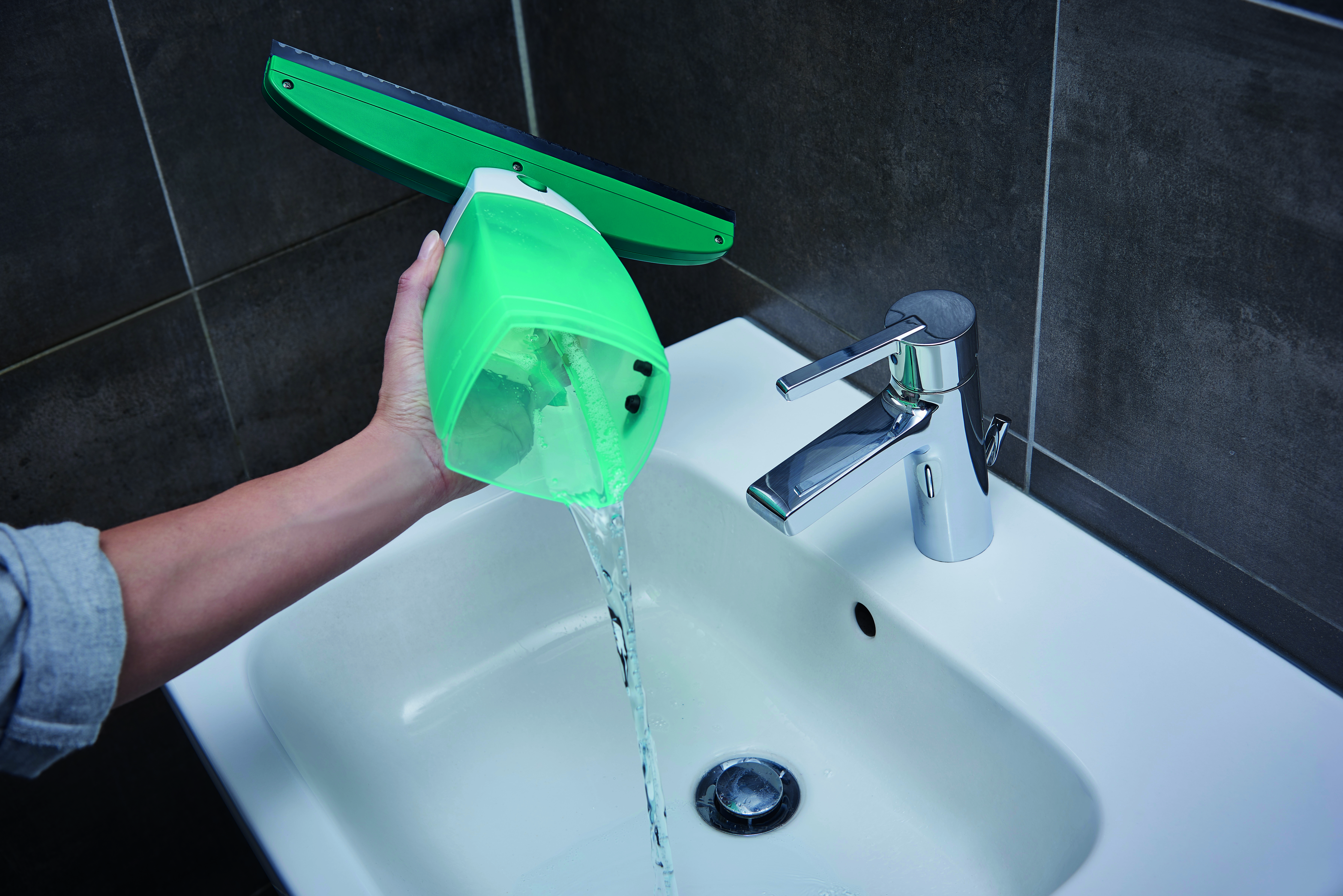 Person füllt grüne Sprühflasche mit Wasser über einem weißen Waschbecken.