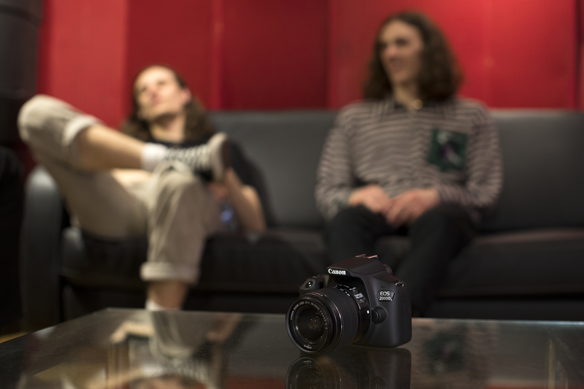 Eine Canon-Kamera auf einem Glastisch, im Hintergrund unscharf zwei Personen auf einem schwarzen Sofa.