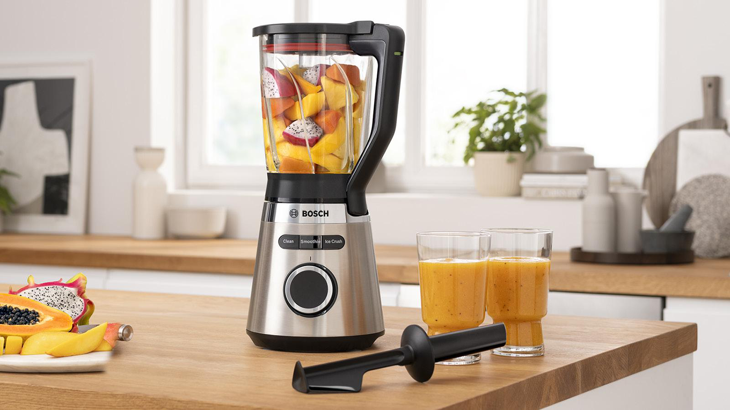 Blender with fruit inside and two glasses of juice. A chopping board is in the foreground.