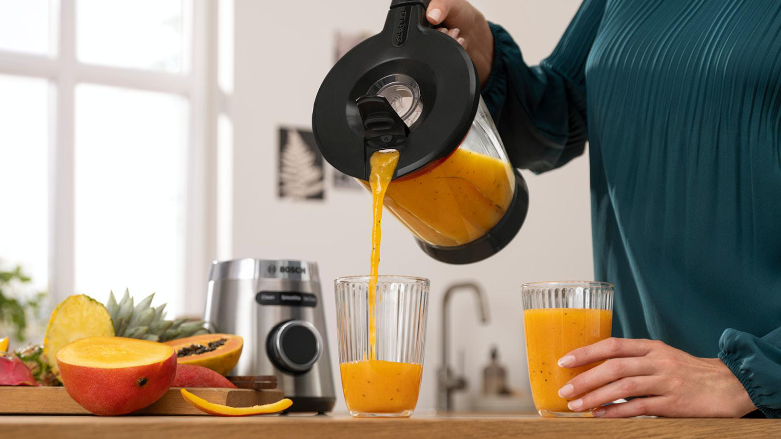 A woman pours orange smoothie from a blender into a glass, with fruits on a table.
