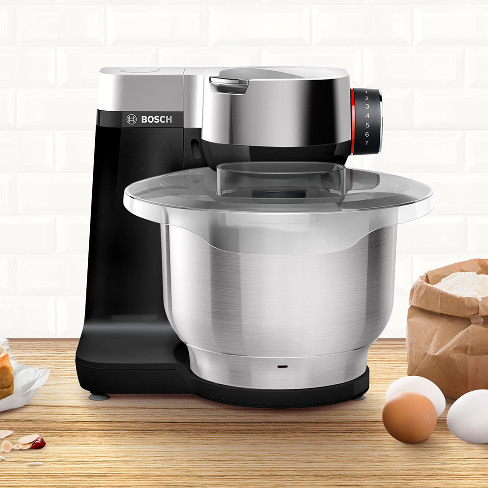 Een zwarte en zilveren keukenmixer met een kom, ingrediënten en een kookboek op een houten tafel.