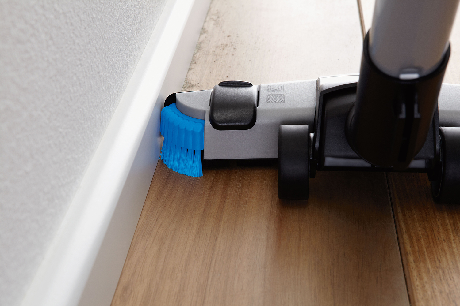 Vacuum cleaner head with blue bristles cleaning a wooden floor edge.