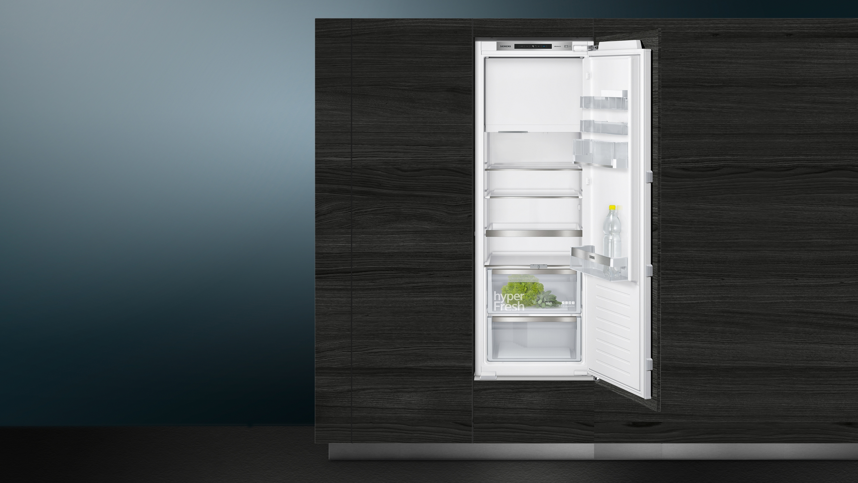 Ein Einbaukühlschrank mit offener Tür, Regalen und einer Schublade mit Gemüse. Dunkle Holzverkleidung.