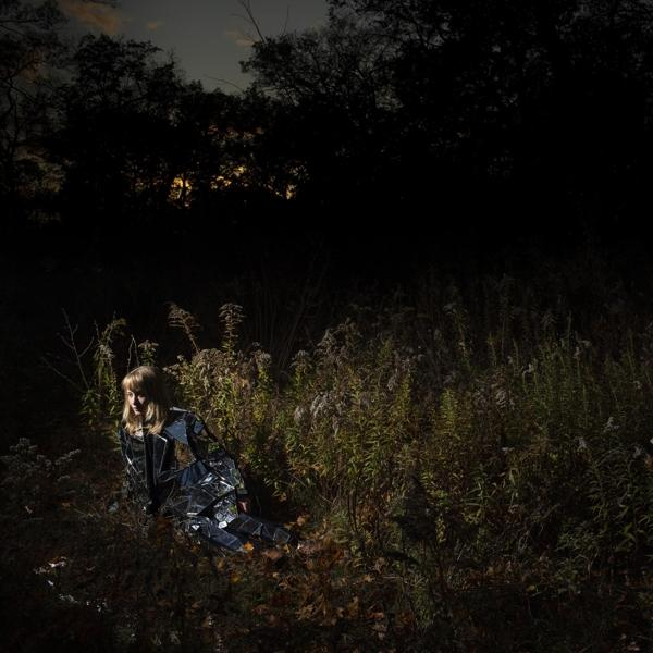Personne dans un champ, portant des vêtements miroir, assise dans les hautes herbes. Ambiance sombre.