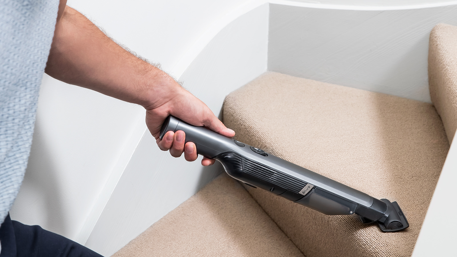 Eine Person benutzt einen grauen Handstaubsauger auf einer beigefarbenen Teppichtreppe. Arm und Hand der Person sind sichtbar.