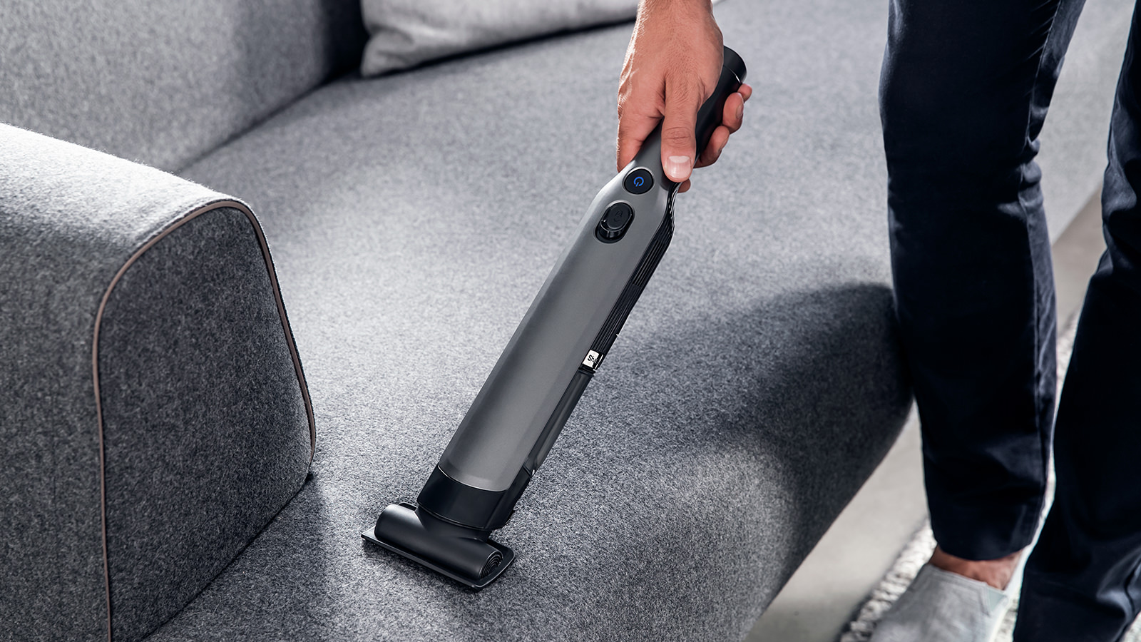 Eine Person saugt mit einem Handstaubsauger ein graues Sofa ab. Der Staubsauger ist dunkelgrau, die Person trägt eine dunkle Hose.