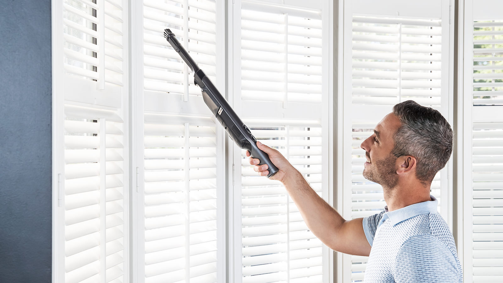 Mann reinigt weiße Fensterläden mit Staubsaugeraufsatz.