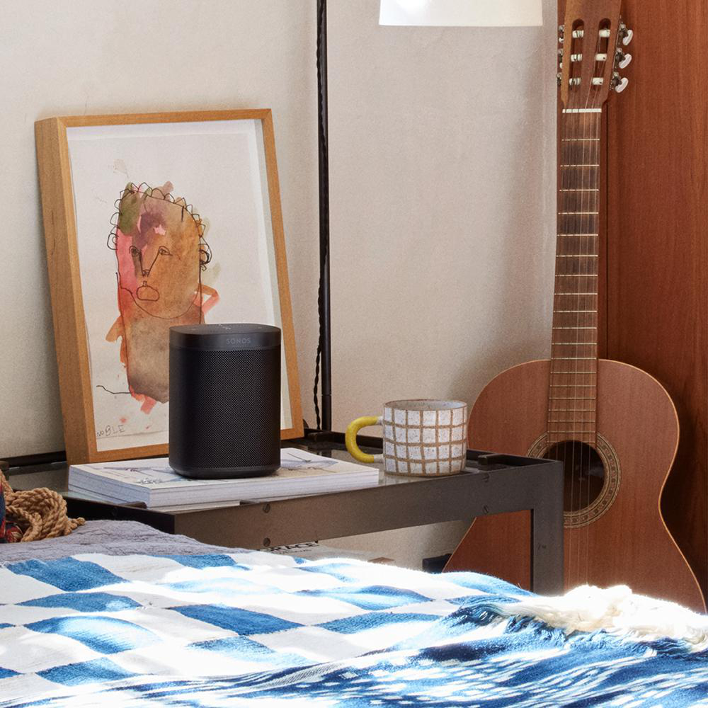 Ein schwarzer Lautsprecher steht auf einem Buch mit Kunst und einer Tasse auf einem Tisch neben einer Gitarre.