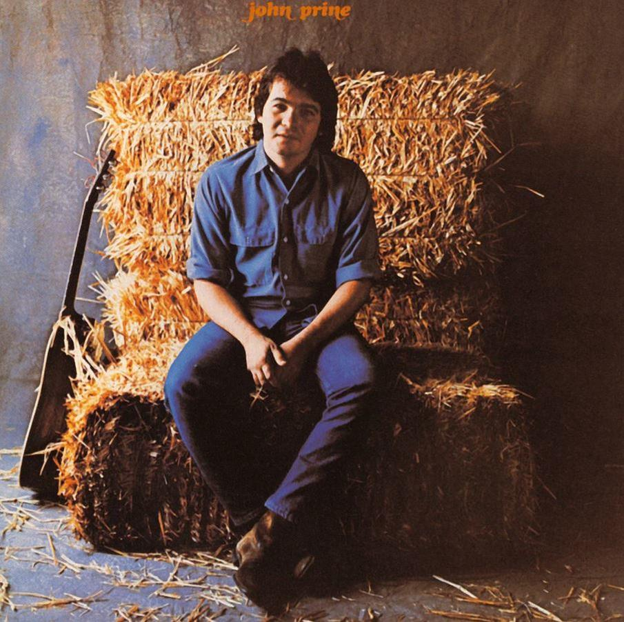John Prine szénabálákon ül, kék ingben és farmerben, gitárral a közelben. Barna és kék tónusok dominálnak.