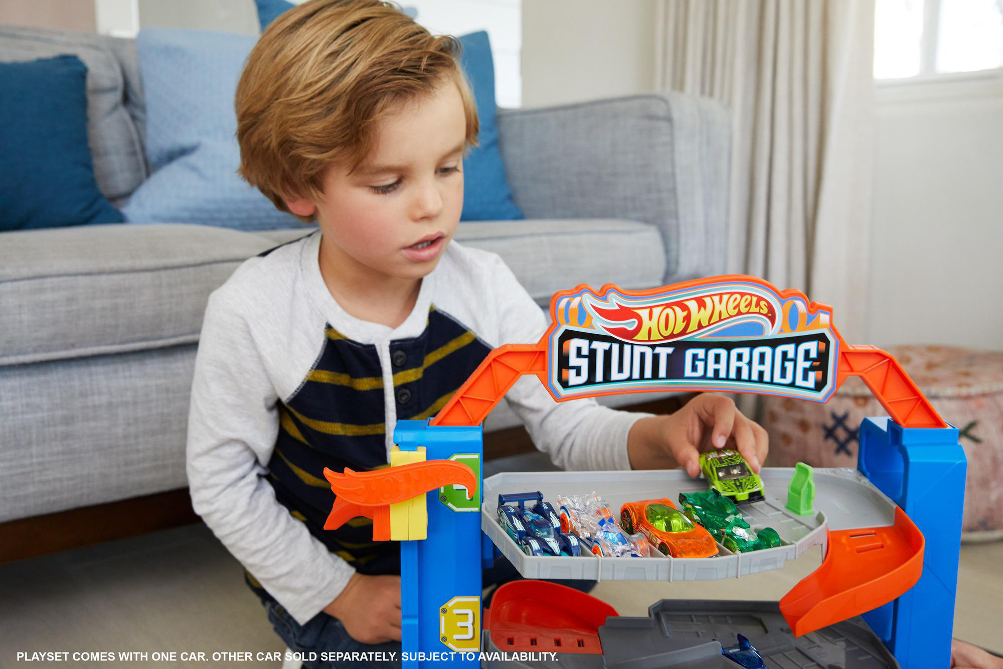Ein Junge spielt mit einer Hot Wheels Stunt Garage. Es gibt Spielzeugautos und eine blau-orangefarbene Strecke. Im Hintergrund ist ein Sofa.