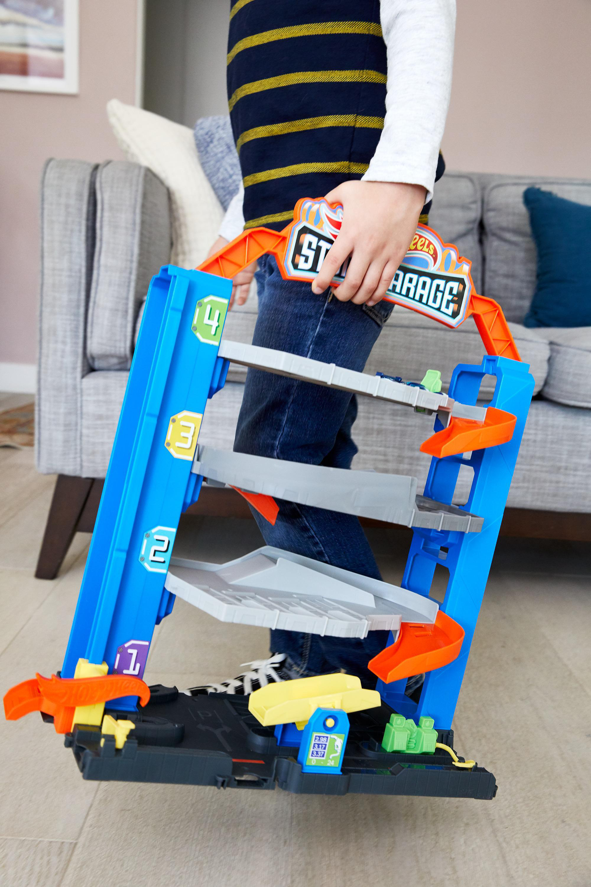 Ein Kind hält eine blau-orangefarbene Spielzeuggarage. Das Spielzeug hat Rampen und Ebenen. Im Hintergrund ist ein graues Sofa.