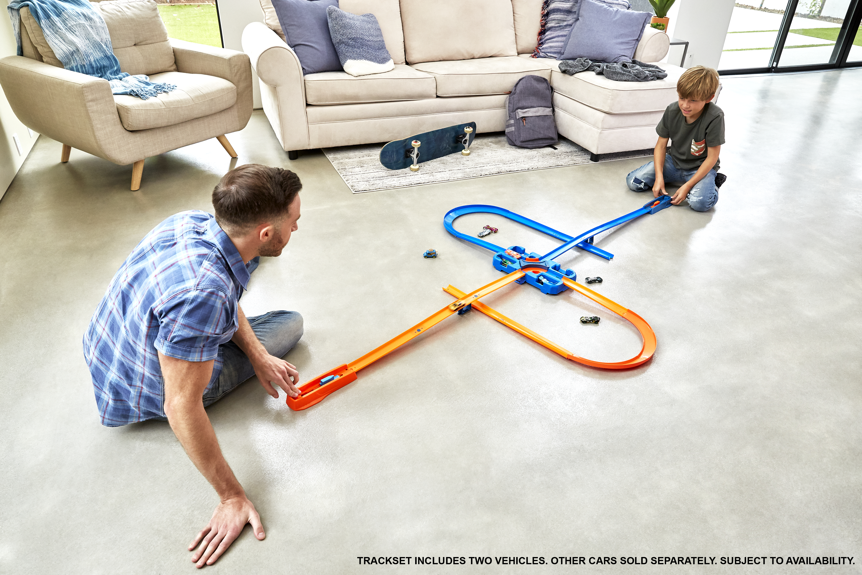 Ein Mann und ein Junge spielen mit Spielzeugautos auf einer Rennstrecke. Im Hintergrund sind ein Sofa, ein Stuhl und ein Skateboard.