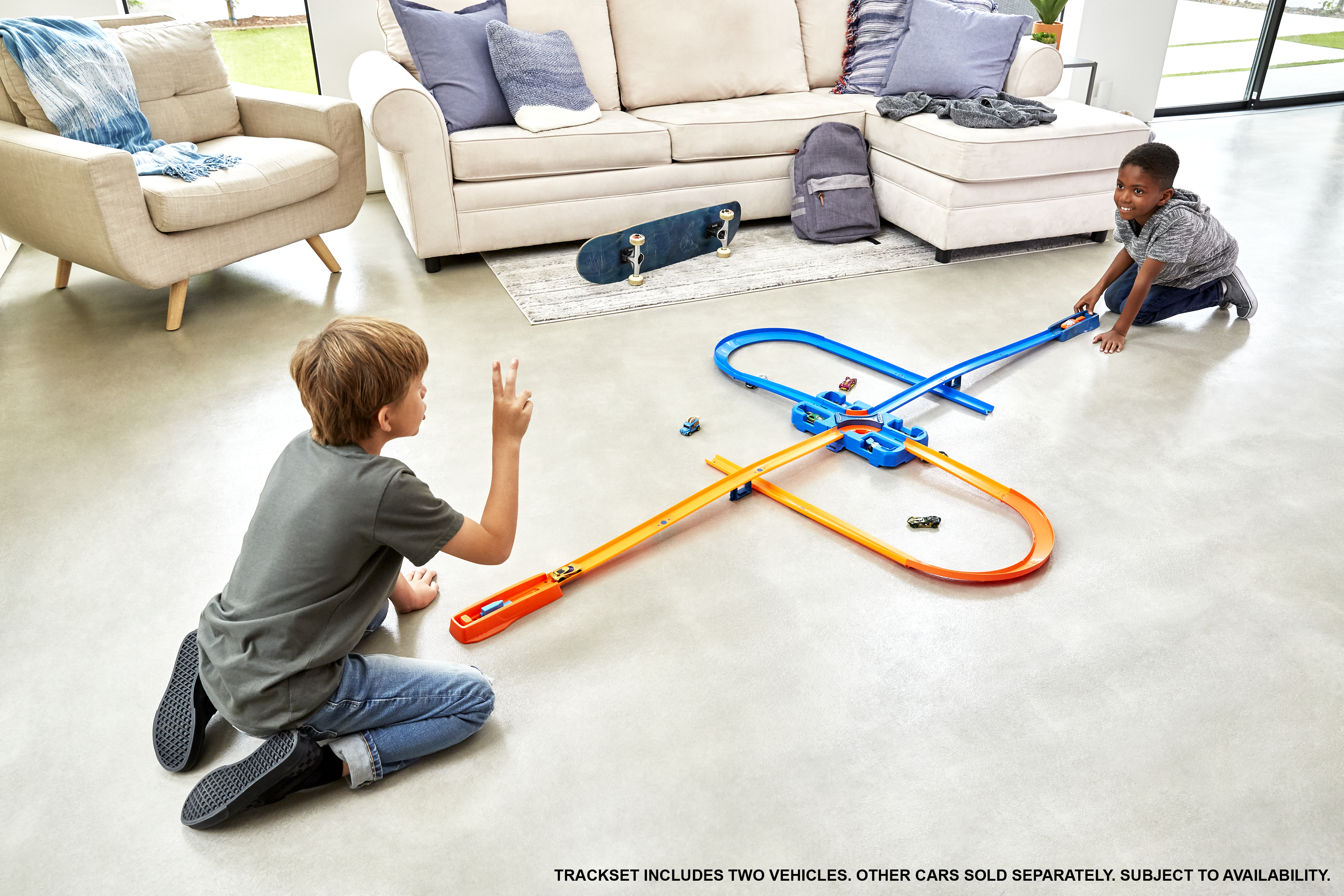 Zwei Jungen spielen mit einer Spielzeugauto-Rennstrecke. Ein Skateboard und ein Rucksack sind sichtbar. Im Hintergrund sind ein Sofa und ein Sessel zu sehen.