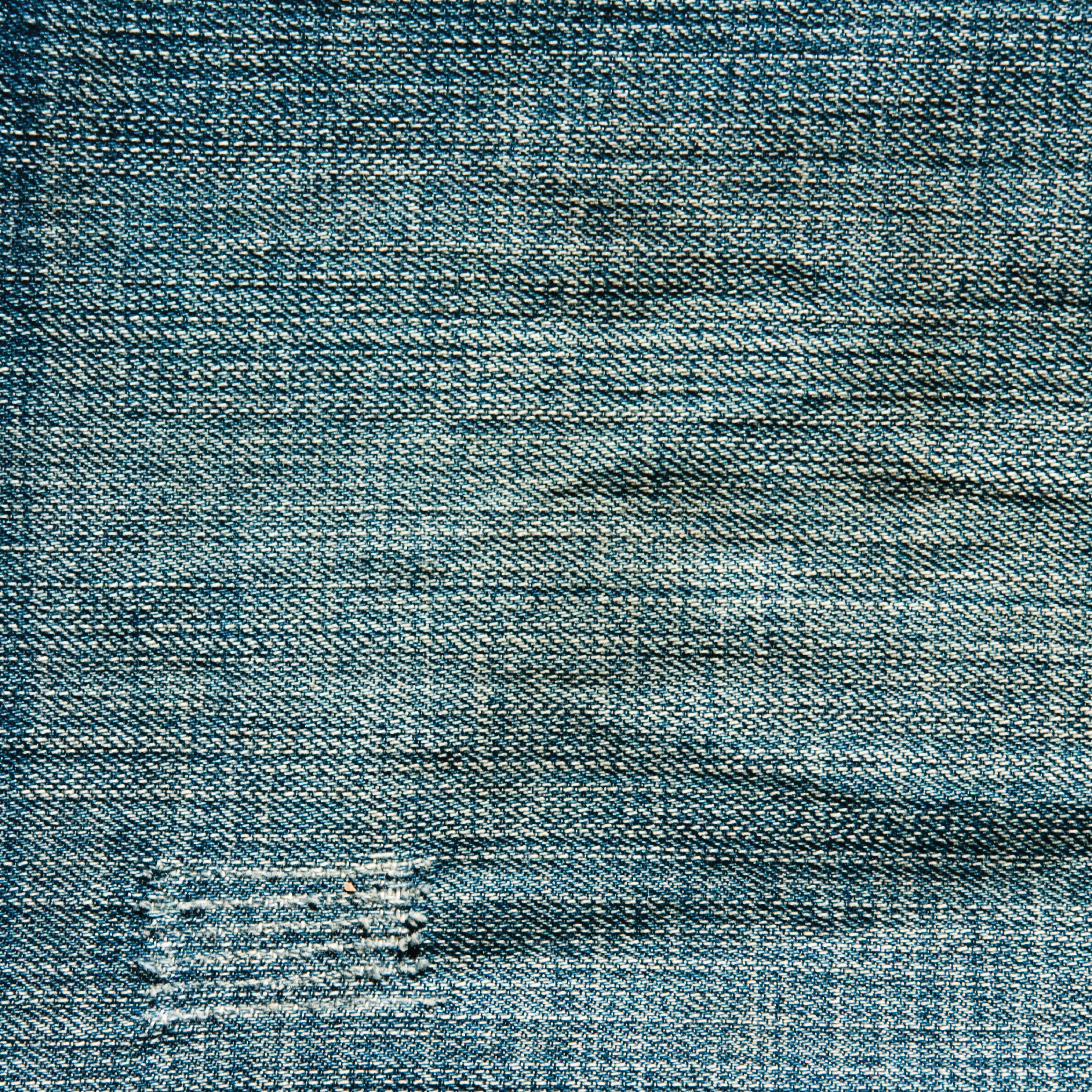 Nahaufnahme von blauem Jeansstoff. Sichtbare Webstruktur mit einem beschädigten Flicken. Ausgeblichene Bereiche und leichte Falten vorhanden.