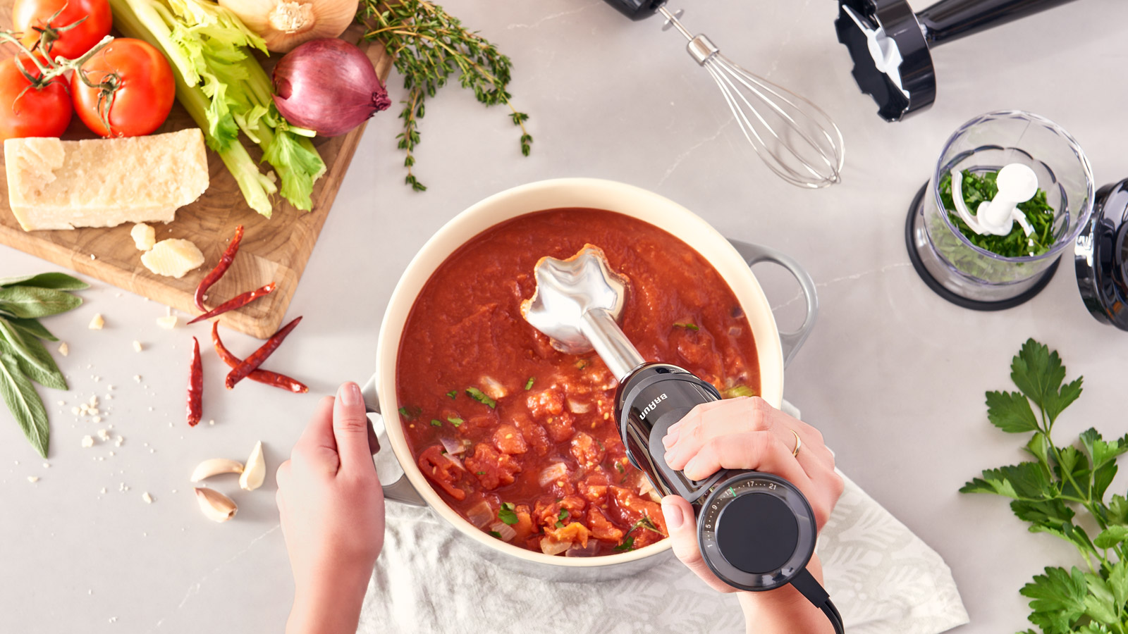 Handen die een blender boven tomatensoep in een grijze pot houden. Ingrediënten eromheen.