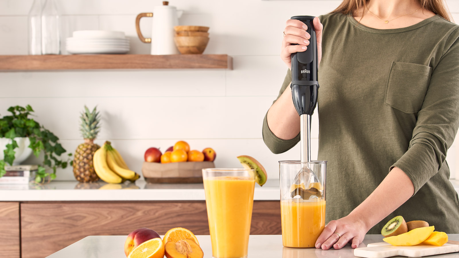 Vrouw gebruikt een zwarte staafmixer in een glas. Fruit op een aanrecht op de achtergrond.