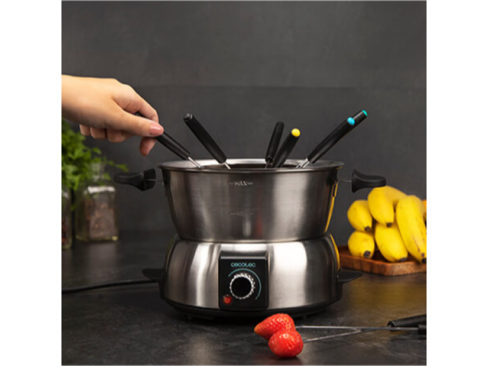 Una olla de fondue con tenedores, una mano que alcanza con un tenedor y algunos plátanos y fresas.