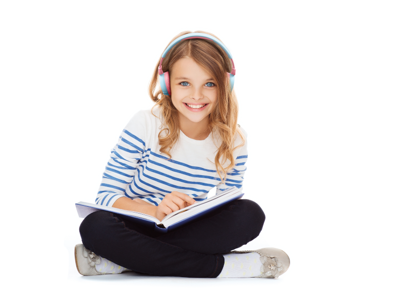 Young girl with headphones smiling, reading a book while sitting with legs crossed, on a white background.