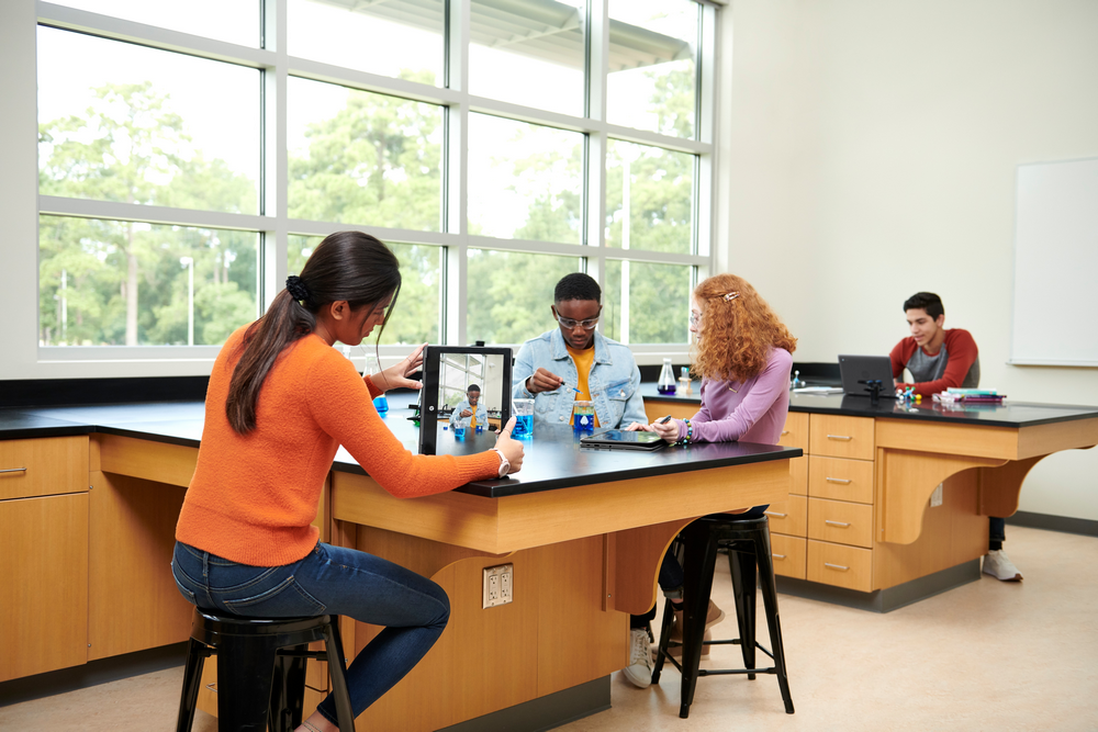 Estudiantes en un laboratorio, usando tabletas y un dispositivo. Mesas, ventanas y vista de árboles al fondo.