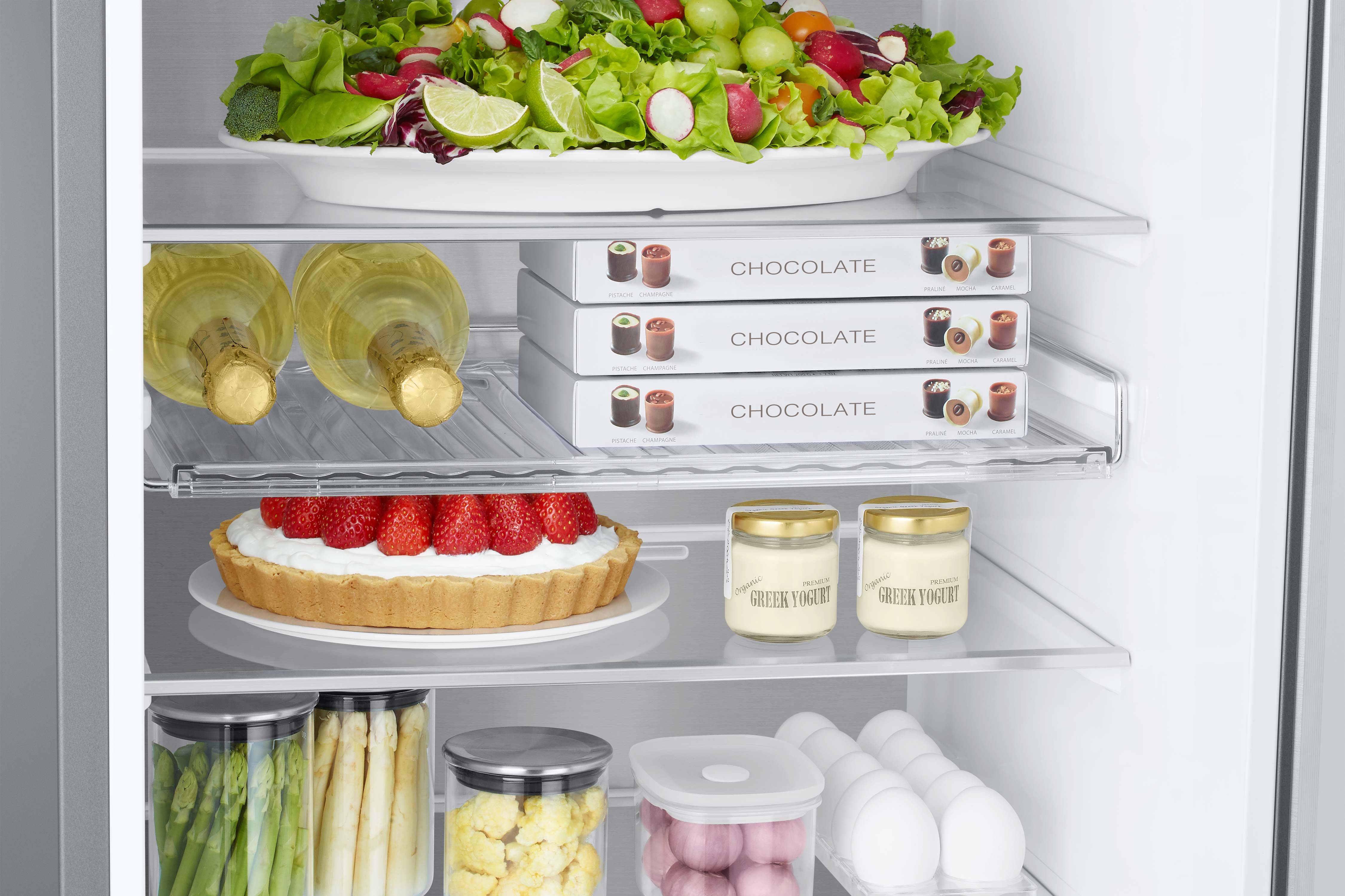 Offener Kühlschrank mit Lebensmitteln: Weinflaschen, Pralinenschachteln, eine Tarte und Joghurt. Salat im obersten Regal.