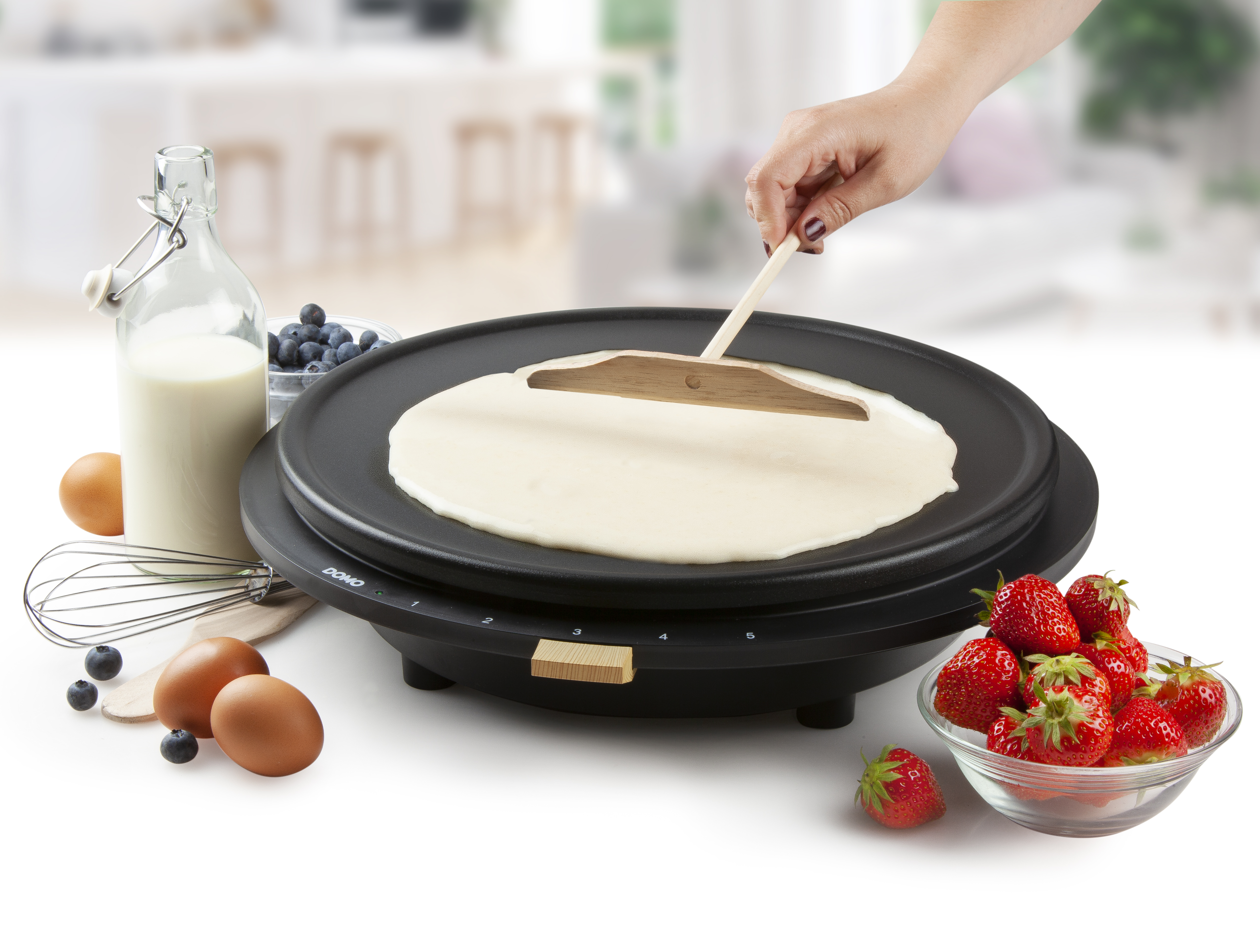 Woman making a pancake on a griddle. Berries, eggs, and milk are nearby.