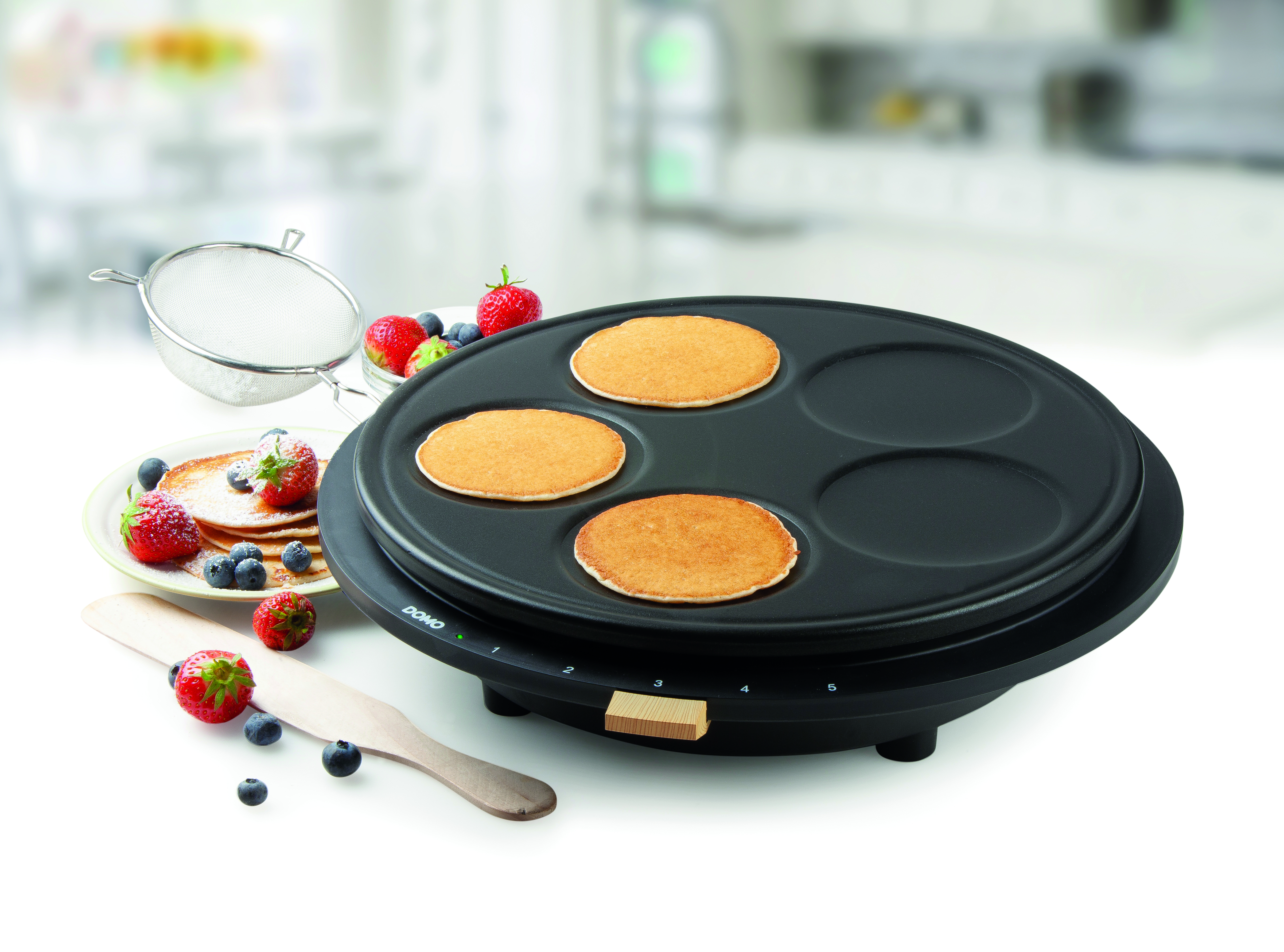 Pancake maker with pancakes and toppings on a white surface.