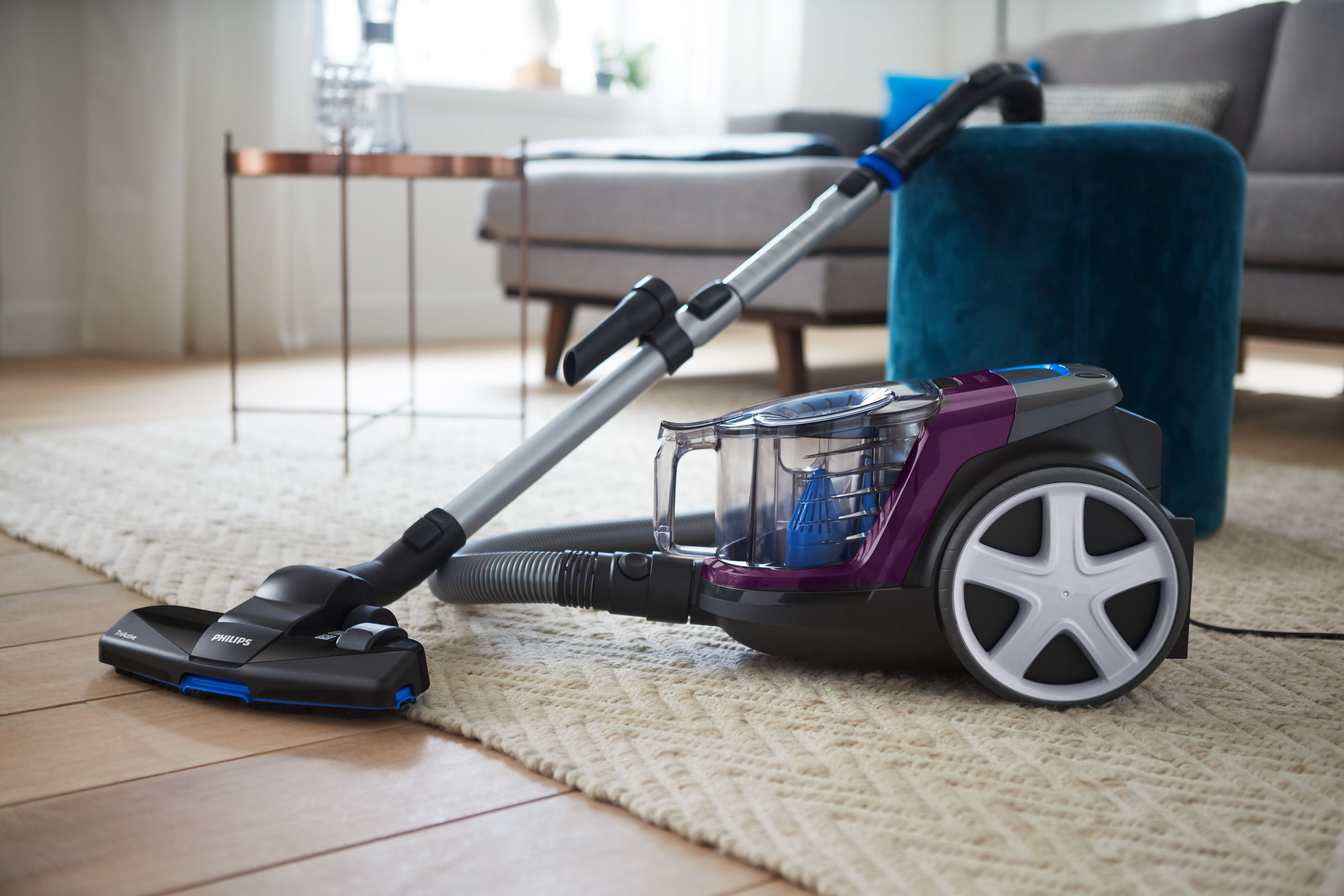 A purple vacuum cleaner is on a rug. Blue and grey furniture is in the background.