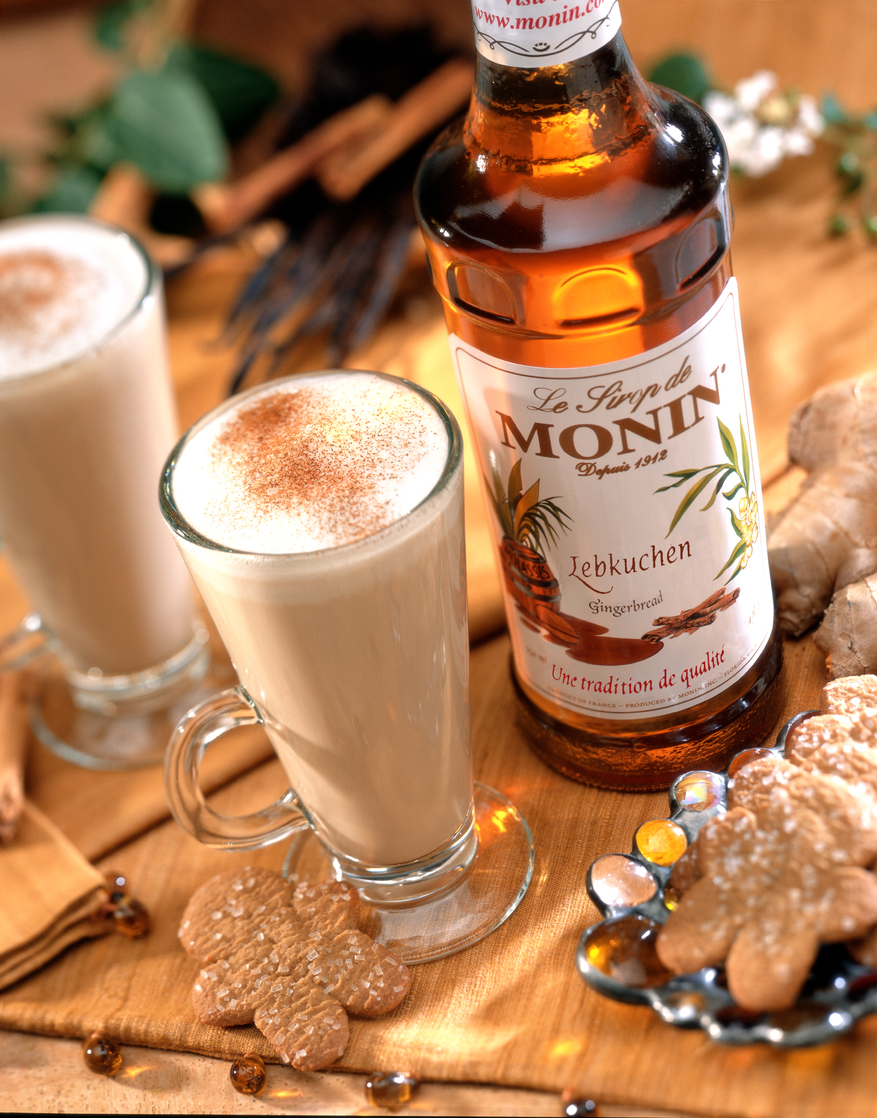Zwei Kaffeegetränke mit Schaum und Zimt, mit Lebkuchen-Sirup-Flasche und Keksen.