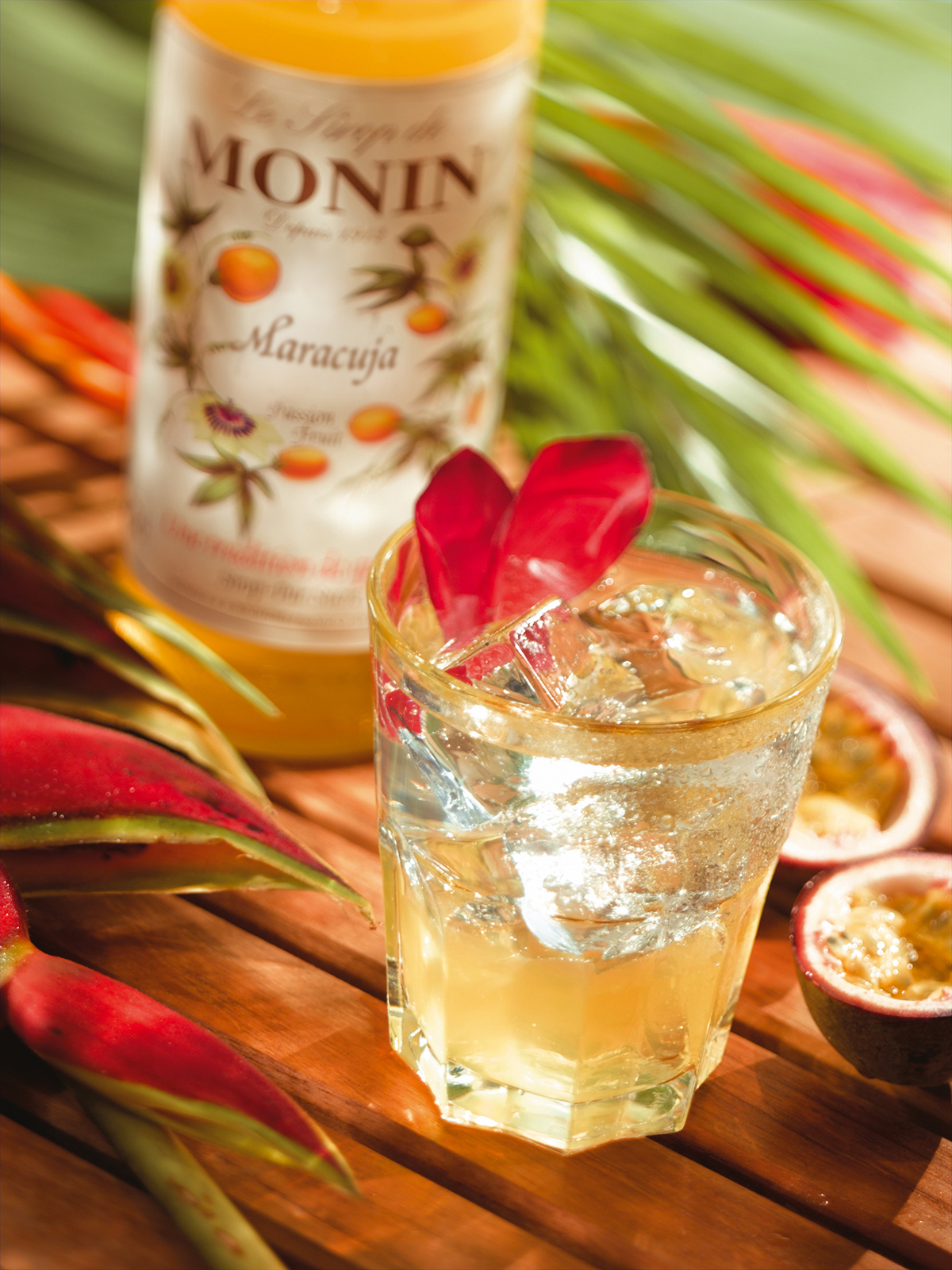 Ein Cocktail mit Eis und einer Passionsfrucht-Garnierung neben einer Monin-Flasche und Passionsfrucht-Hälften auf Holz.