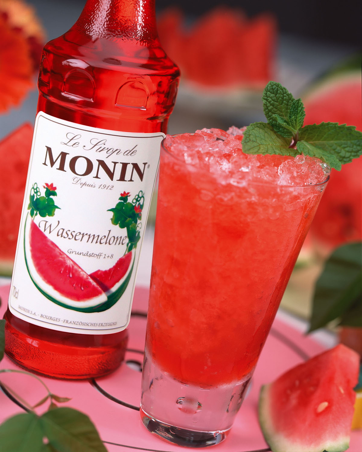Ein Wassermelonengetränk mit Eis und Minze. Eine Sirupflasche steht neben dem Glas. Wassermelonenscheiben im Hintergrund.