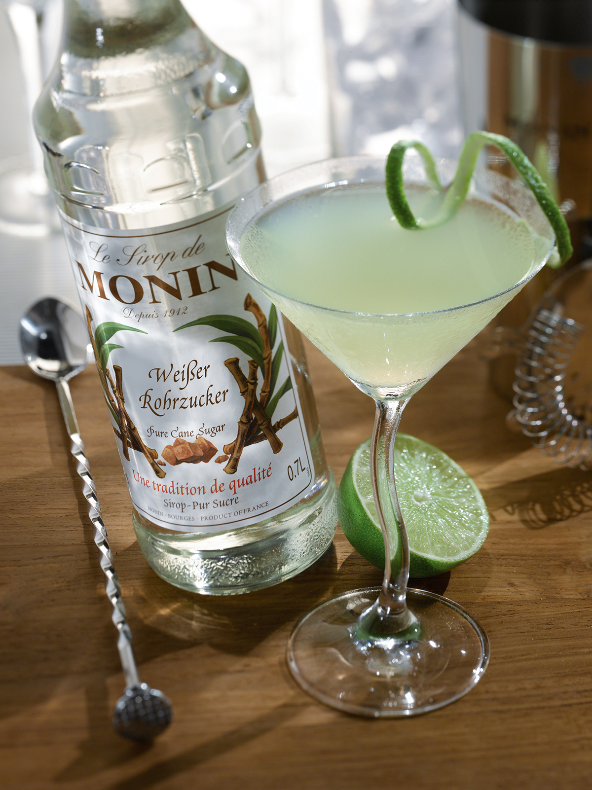 Cocktail im Martini-Glas, Limettenhälfte, Monin-Sirupflasche und Barlöffel auf Holzoberfläche.