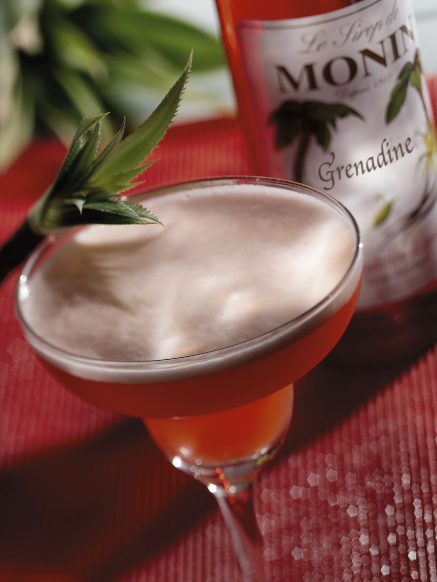 Ein Cocktail in einem runden Glas mit einer Flasche Grenadine-Sirup.