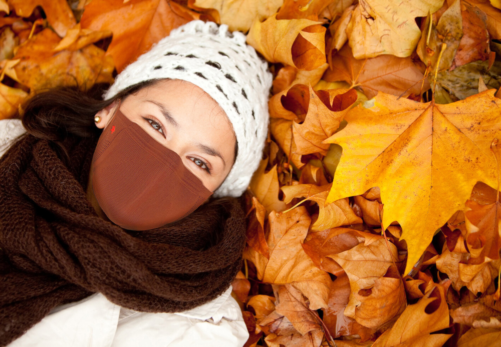 Een vrouw met een witte hoed, bruine sjaal en bruin gezichtsmasker, liggend op herfstbladeren.